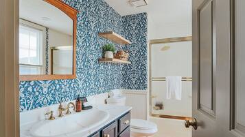 A bathroom with a white sink, gold faucets, and a blue patterned wall. There are two wooden shelves above the sink, one holding a plant and the other a decorative item.
