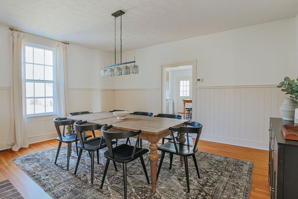 A dining room with a wooden table, six chairs, a chandelier, and a large window with curtains.