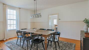 A dining room with a wooden table, six chairs, a chandelier, and a large window with curtains.