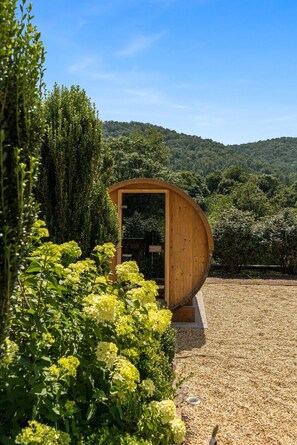 A wooden structure with a curved roof, surrounded by greenery and a gravel path.