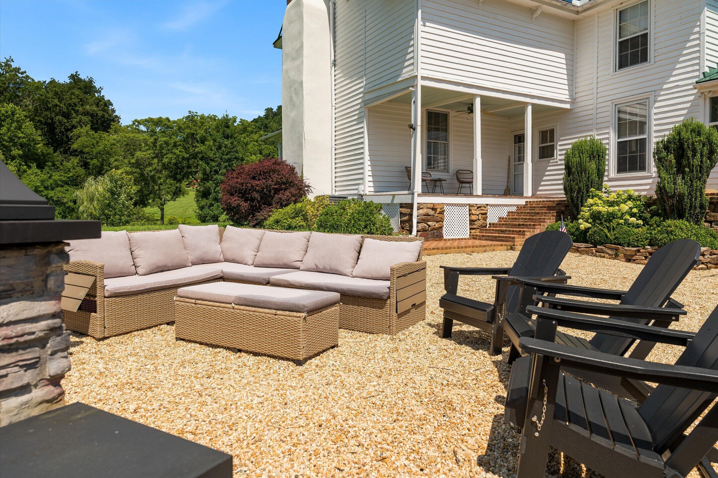 A covered outdoor seating area with a sectional sofa and Adirondack chairs, a stone wall, and a house with a porch.
