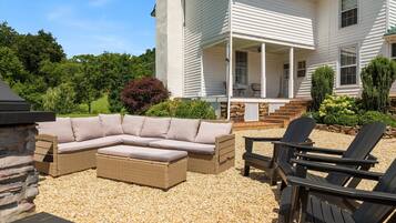 A covered outdoor seating area with a sectional sofa and Adirondack chairs, a stone wall, and a house with a porch.