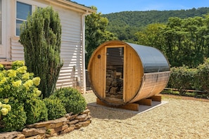 A wooden barrel-shaped structure with a stone interior, situated on a gravel surface, adjacent to a house with a white exterior and a lush green landscape in the background.