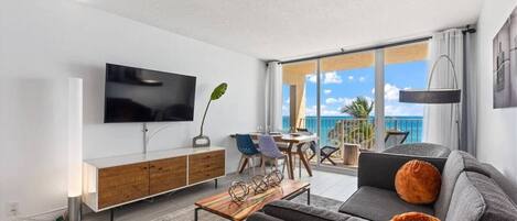 Ein modernes Wohnzimmer mit Flachbildfernseher, Holzschrank, grauem Sofa und einem Balkon mit Meerblick.