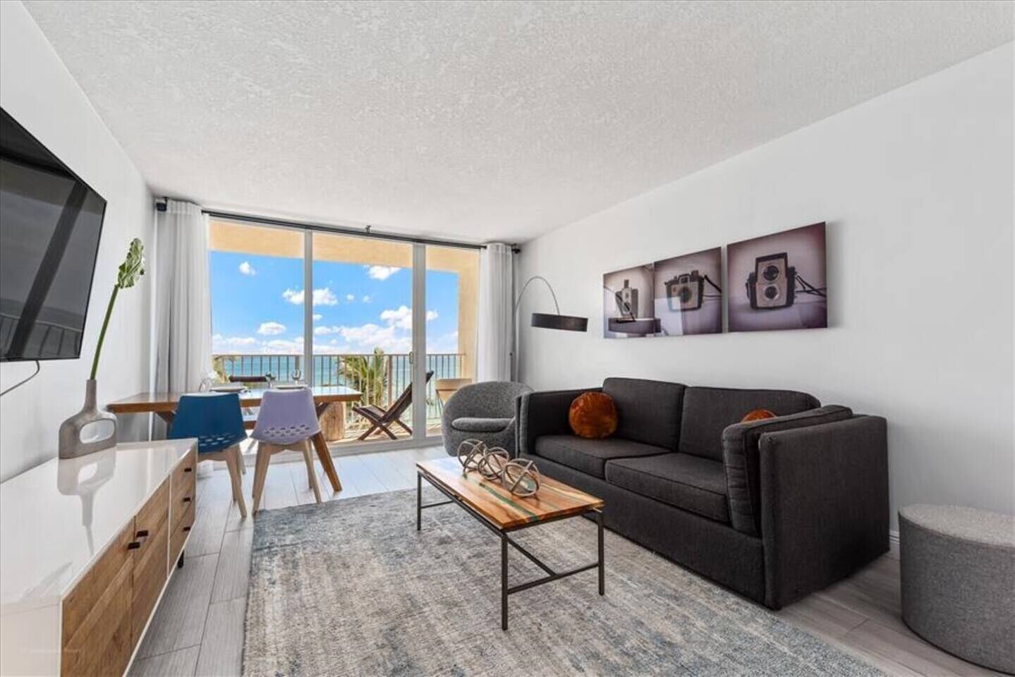 A modern living room with a grey sofa, a wooden coffee table, and a balcony with a view of the sea.