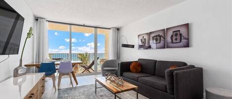 A modern living room with a grey sofa, a wooden coffee table, and a balcony with a view of the sea.