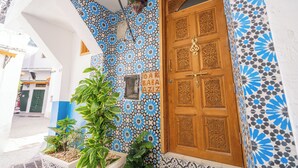 Een gebouw ingang met een muur in blauw en wit patroon, een houten deur met ingewikkelde snijwerk, en een bord met de tekst 'DAR BABA AZIZ'.