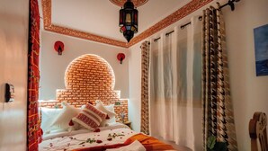 Een hotelkamer met een hoofdbord met een baksteenmotief, een bed met witte lakens en rozenblaadjes, een hanglamp en gordijnen met een patroon.
