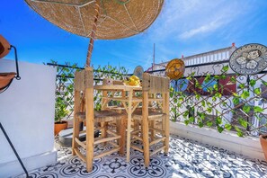 A wooden table and chairs with a woven umbrella, potted plants, and decorative tiles on a balcony.