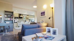 Un salón moderno con un sofá gris, una mesa de centro blanca con revistas, una estantería y una cocina al fondo.