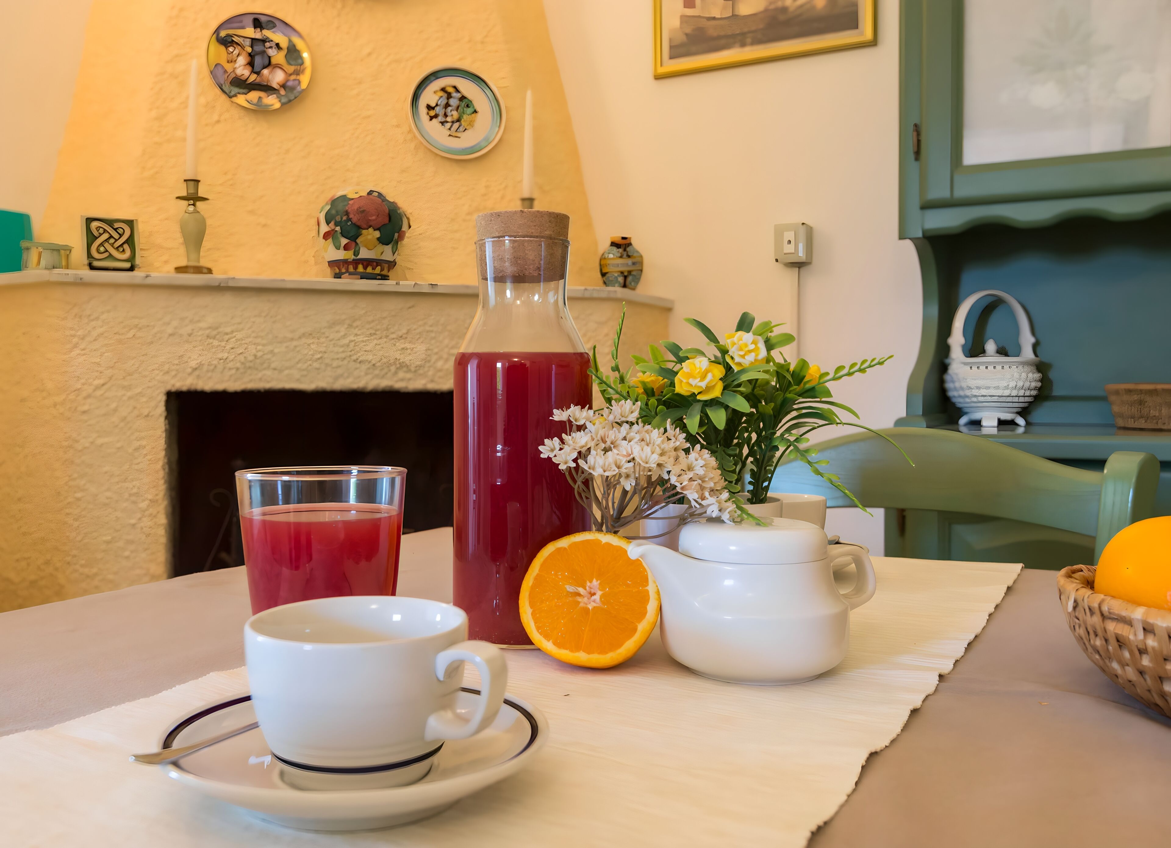 Una mesa de comedor con una taza blanca, un vaso de bebida roja, una botella de bebida roja, media naranja, una jarra blanca y un jarrón con flores.