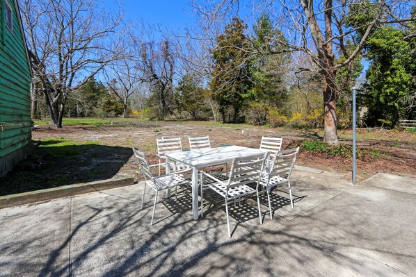 A patio with a table and chairs, surrounded by trees and a green building. - Charming 3-bedroom farmhouse with AC, WiFi, 5 minutes to Sea Isle Beaches! (Dennis)