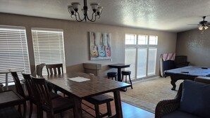 A spacious living room with a wooden dining table, comfortable seating, a pool table, and large windows with blinds.