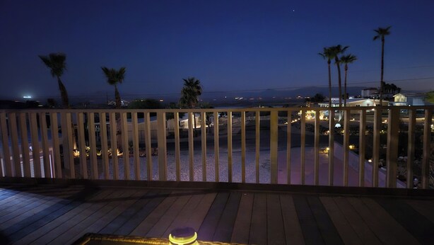 A wooden deck with a table and a lamp, overlooking a pool area with palm trees and distant buildings.