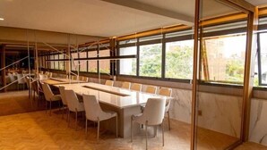 A conference room with a long table, white chairs, and large windows offering a view of the outdoors. - Charming 1-bedroom apartment with AC, WiFi in peaceful Bela Vista Porto Alegre (Rio Branco Porto Alegre)