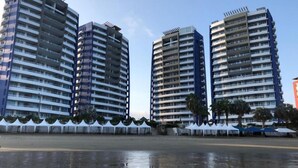 Vista al agua de edificios altos con balcones, una playa de arena y carpas blancas a lo largo de la orilla.