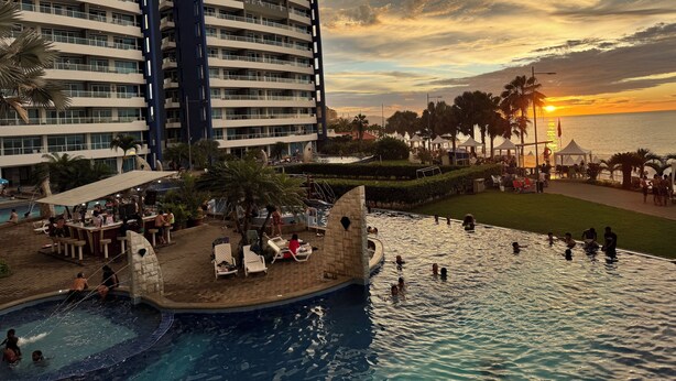 Zona de piscina en un resort con vistas a la puesta de sol, palmeras y un gran edificio al fondo.