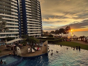 A resort pool area with a view of the sunset, palm trees, and a large building in the background.