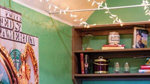 Eine Barbereich mit grüner Wand, einer Holztheke und schwarzen, gepolsterten Barhocker. An der Wand hängt ein Poster und auf der Theke steht eine Lavalampe.