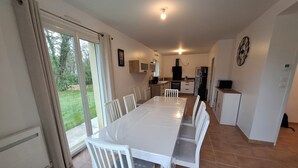 Un comedor con una mesa larga y sillas, una cocina al fondo y vista a zonas verdes exteriores.