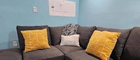 Una sala moderna con un sofá seccional, dos almohadas amarillas, un plaid estampado de cebra y una pared azul claro.