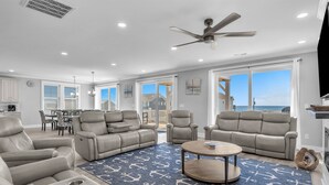 A spacious living room with a ceiling fan, large windows, and a view of the ocean. - Brand New Oceanview 8bed/7.5 bath Hot Tub EV Charger (Surf City)
