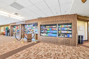 A hotel lobby with an 'ESSENTIALS' kiosk stocked with snacks and beverages, a trash can, and a wooden-patterned carpet.