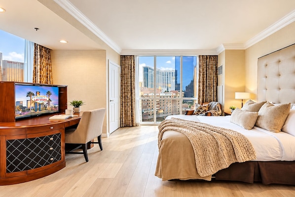 A hotel room with a bed, a desk with a television, a view of the city through large windows, and a balcony.