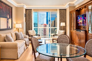 A modern hotel room with a glass table, comfortable seating, a television, and a view of the cityscape.