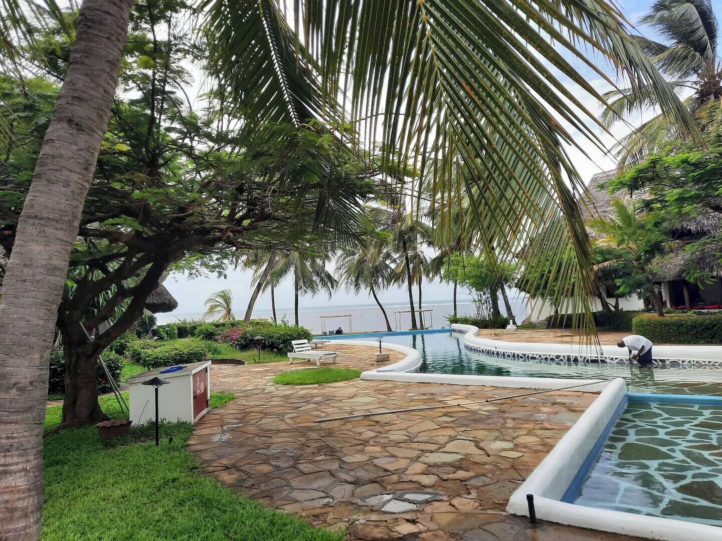 A pool area with stone flooring, palm trees, and a view of the sea.