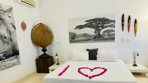 Une chambre d’hôtel avec un lit, un climatiseur fixé au mur, une photographie encadrée, des objets de décoration et un arrangement floral en forme de cœur posé sur le lit.