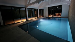 Una piscina interior con techo de cristal, suelo de baldosas y zona de estar con sillas y una mesa.