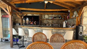 An outdoor bar area with a stone wall, wooden ceiling, and a TV screen displaying a fire. There are wicker chairs and a table with a bar counter.