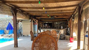 A covered outdoor seating area with wicker chairs and a glass table, a stone wall with a bar and fireplace, and a pool in the background.
