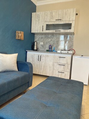 A modern living room with a sofa, a coffee table, a kitchen area with cabinets and appliances, and a wall-mounted light fixture.