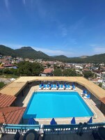 Piscina con sillas de descanso y sombrillas, rodeada por una terraza de madera y vistas a un pueblo y montañas.