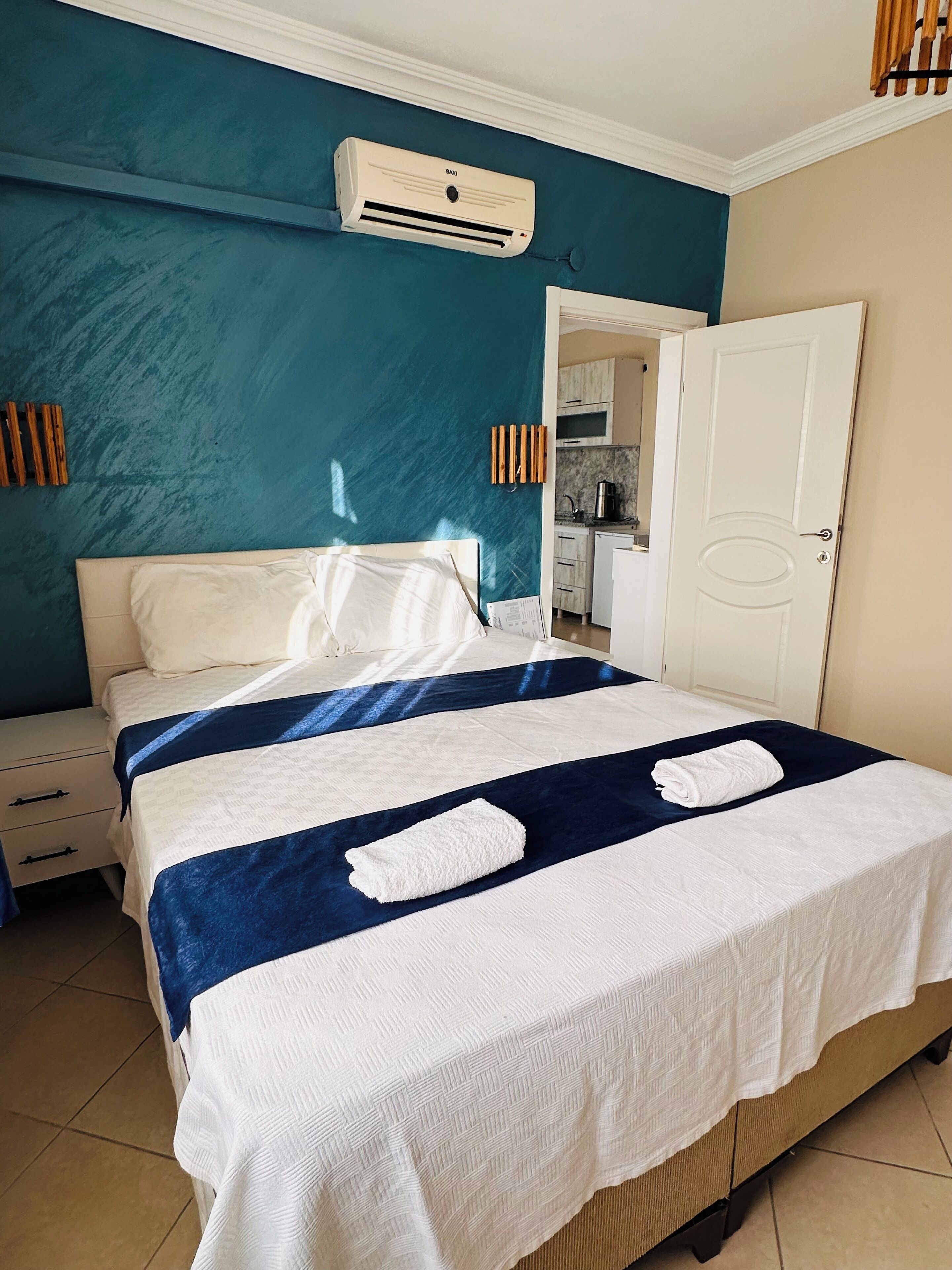 Ein ordentlich bezogenes Bett mit einer weiß-blauen Tagesdecke, einem weißen Kopfkissen und einem weißen Handtuch. Eine weiße Nachttisch mit Schubladen befindet sich links neben dem Bett. Eine weiße Tür ist rechts vom Bett sichtbar.