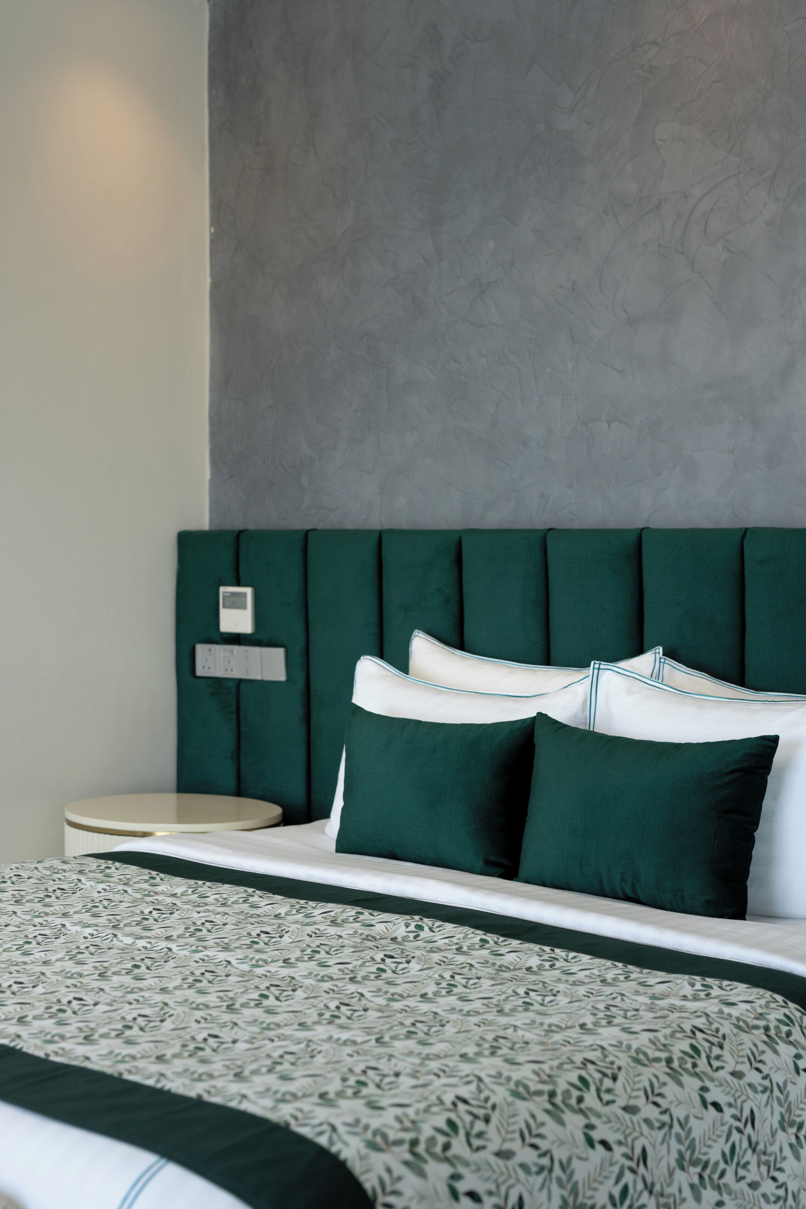 A bed with a patterned bedspread, a green upholstered headboard, and a bedside table with a control panel.
