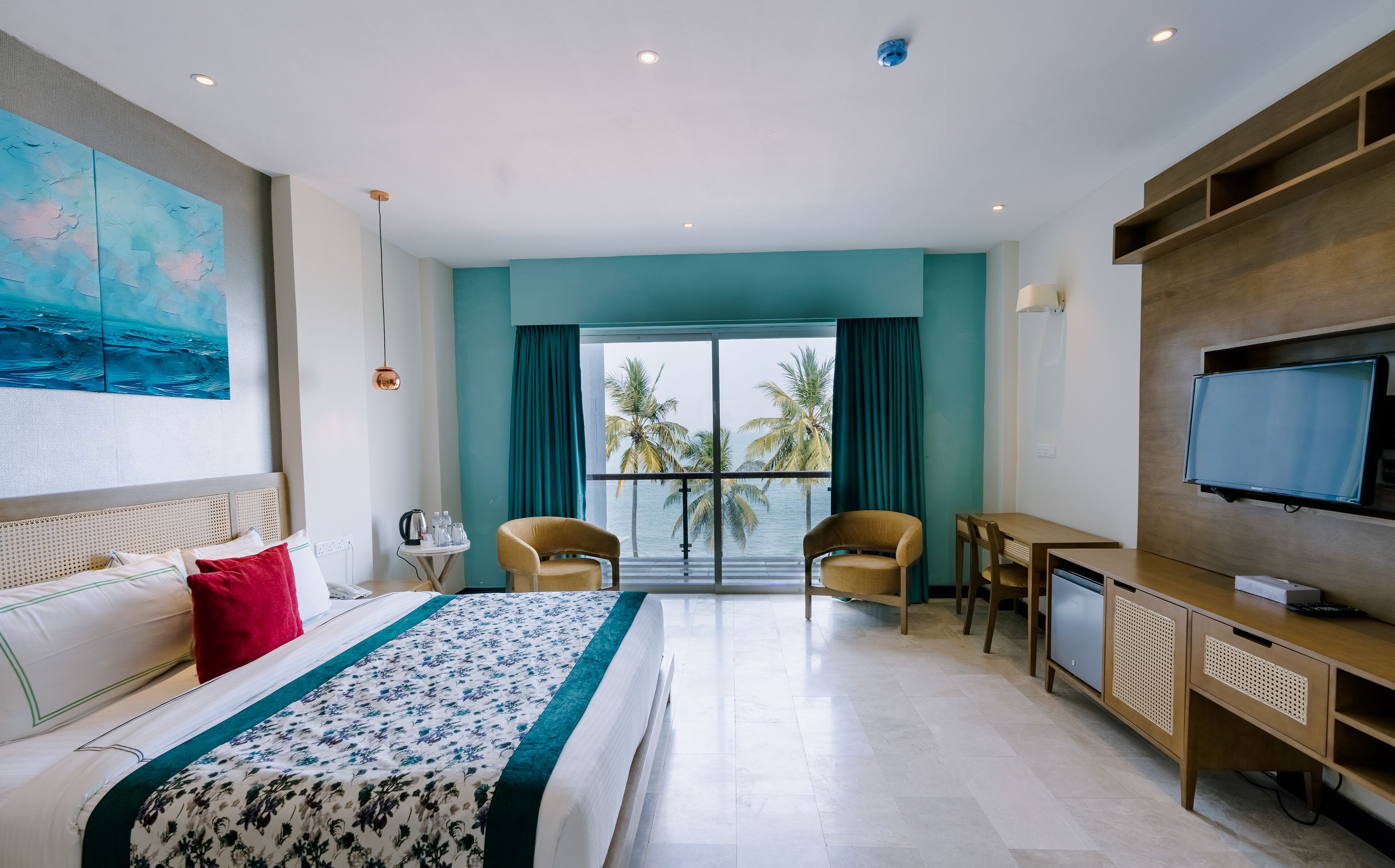 Habitación de hotel con cama, escritorio, televisor y vistas a palmeras y al mar.