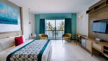 Ein Hotelzimmer mit Bett, Schreibtisch, Fernseher und Blick auf Palmen und das Meer.
