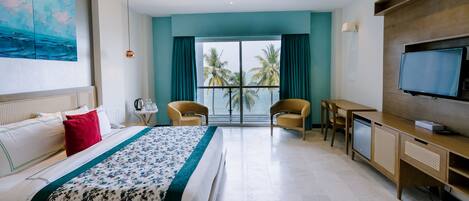 Habitación de hotel con cama, escritorio, televisor y vistas a palmeras y al mar.