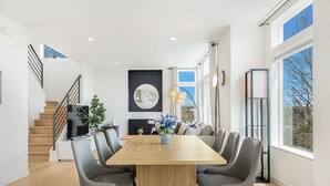 Un espace repas moderne avec une table en bois et des chaises grises, un escalier et une grande fenêtre donnant sur un beau paysage.