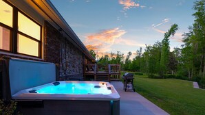 Un bain à remous dans un jardin, avec un mur en pierre, une terrasse en bois et une zone herbeuse agrémentée d’arbres.
