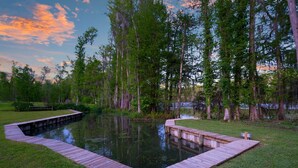 Une terrasse en bois entoure un étang rectangulaire, lequel est traversé par un petit pont en bois. L’ensemble est bordé de grands arbres et d’une zone herbeuse.