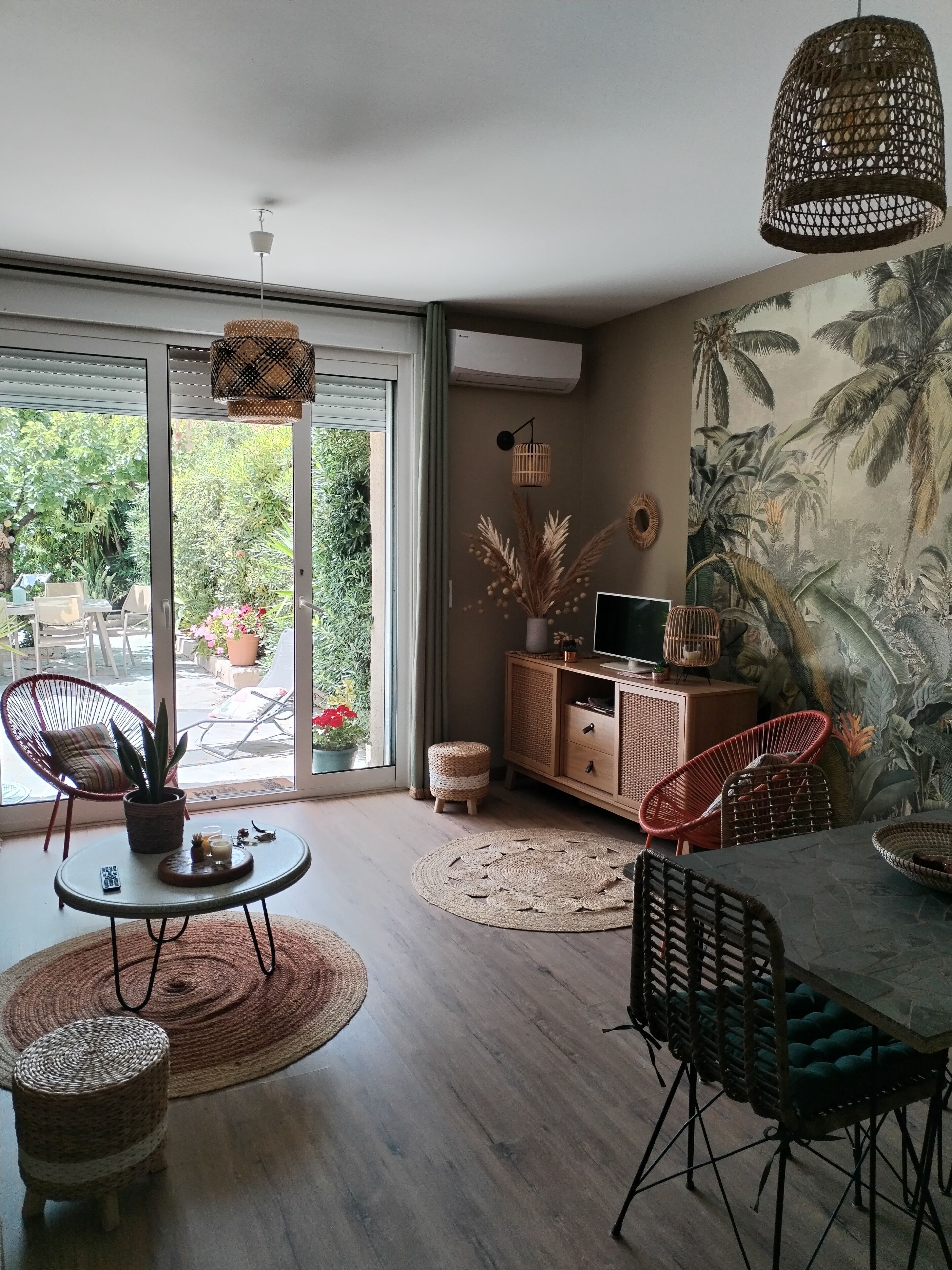 Ein modernes Wohnzimmer mit einem großen Schiebeglastür, die zu einem Garten führt, Korbmöbeln, einer gewebten Pendelleuchte und einem tropischen Wandbild.