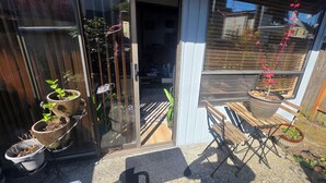 Une porte vitrée donne accès à l’intérieur, avec des plantes en pot sur la gauche, un banc en bois sur la droite, et une petite table avec une chaise devant.