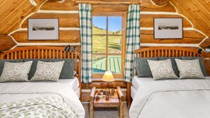 A cozy log cabin bedroom with two beds, a small table with pinecones, and a view of a green landscape through a large window.
