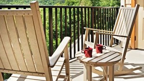 Un balcon avec deux chaises en bois et une table, une rambarde noire et une vue sur une colline verdoyante et boisée.