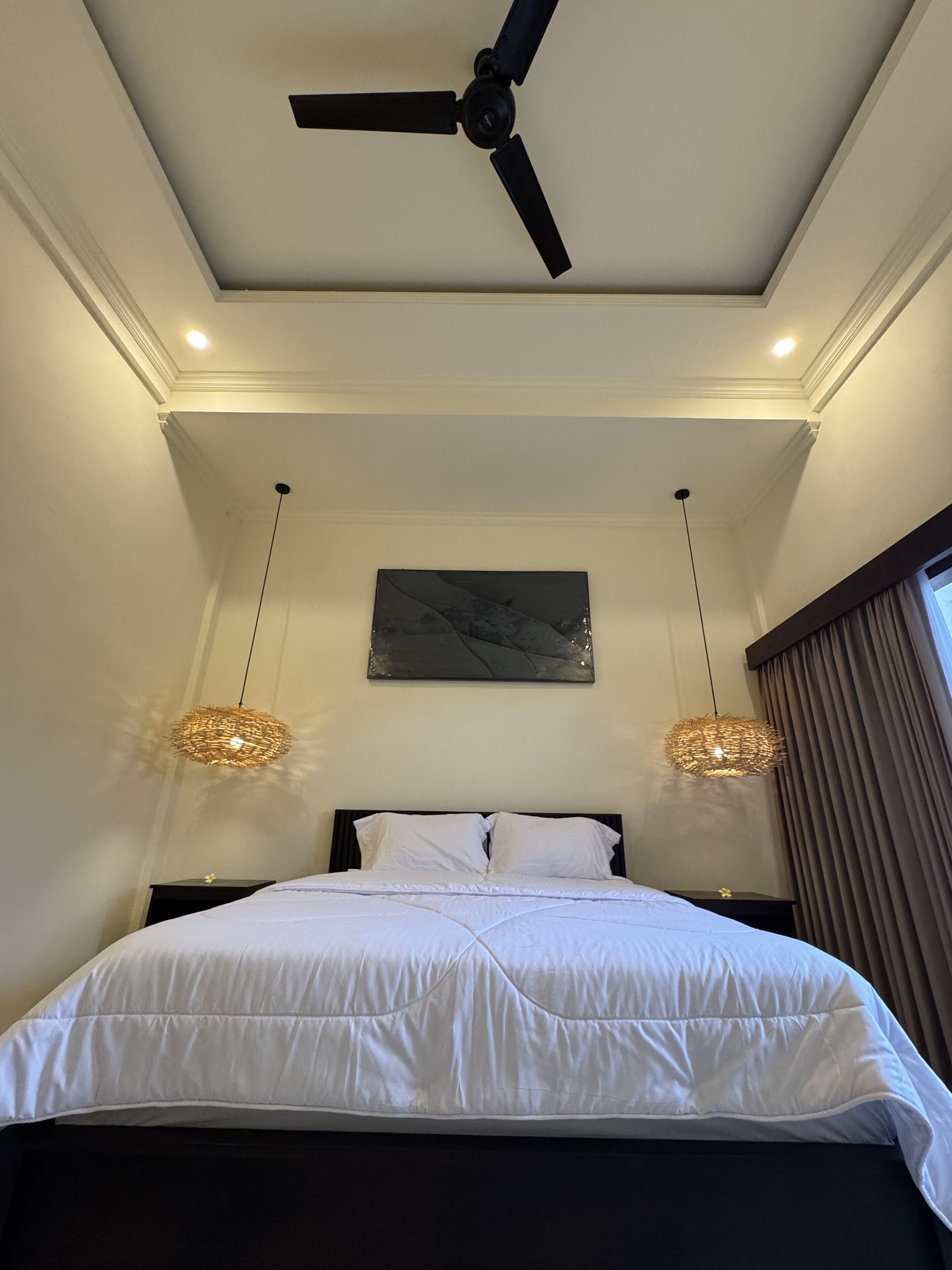 A bedroom with a ceiling fan, two pendant lights, a bed with white bedding, a dark headboard, a wall-mounted artwork, and a window with curtains.