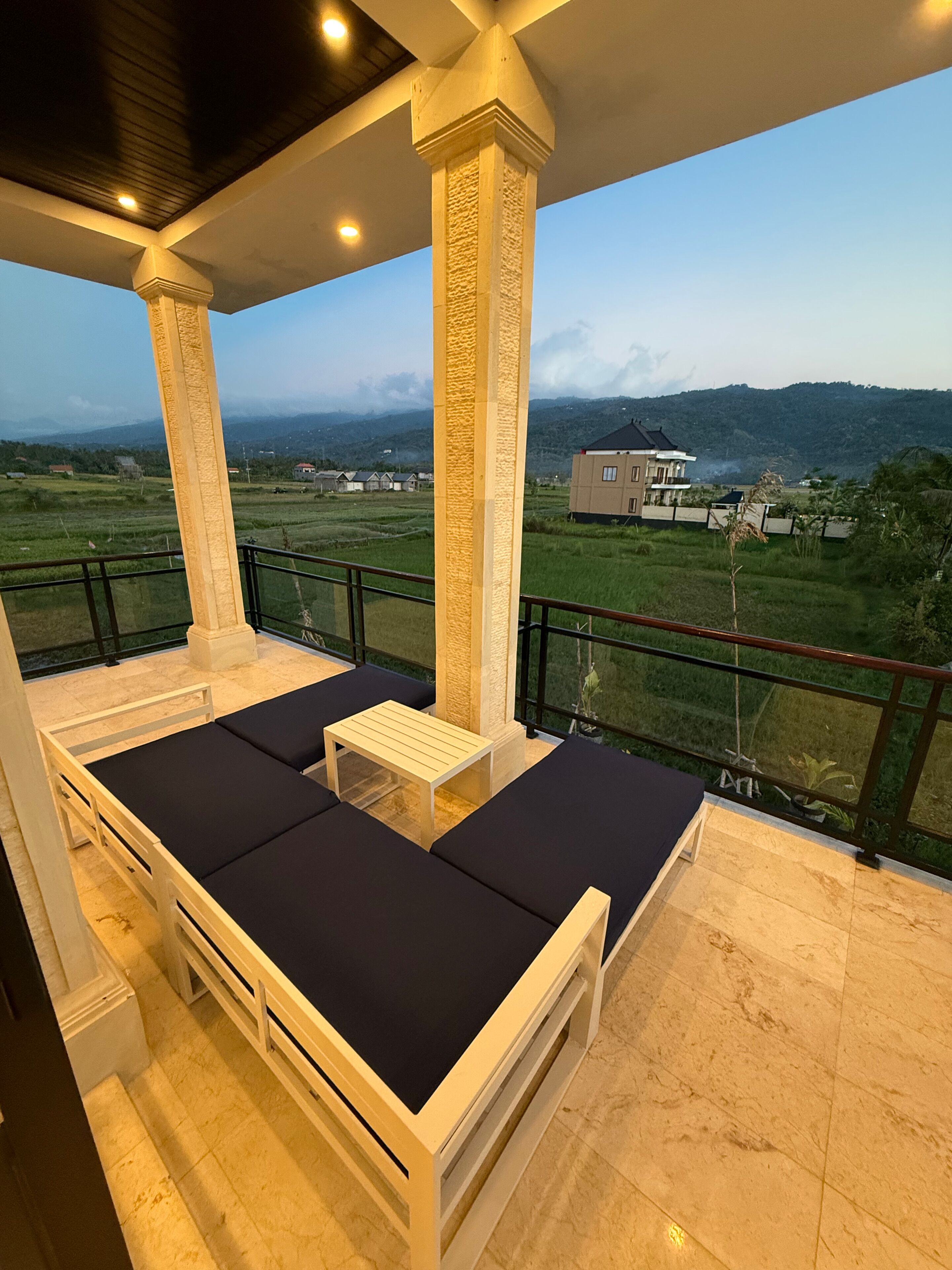 Un balcón con vistas a un paisaje montañoso, una cama blanca con cojines oscuros y una pequeña mesa de madera.